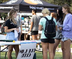 Three young women chat with young woman standing behind a table with a sign that reads Pre-law club.