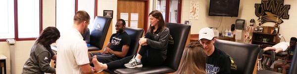 In the athletic training room, Professor Wilson sits while a student wraps her ankle.