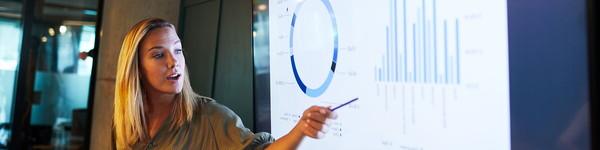 Business woman in front of a digital screen pointing to graphs while speaking.