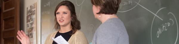 Two females—a student and a professor— stand in a classroom in front of a chalk board.