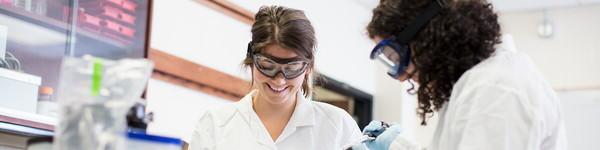 Two female student in scrubs and protective eyewear in a chemistry lab.