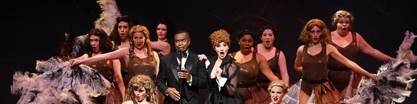 A man in a tux, a woman in a gown and dancers with large feather fans perform on stage.