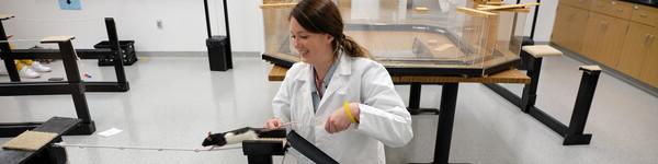 Female student coaches a black and white rat across a tied rope in a lab.