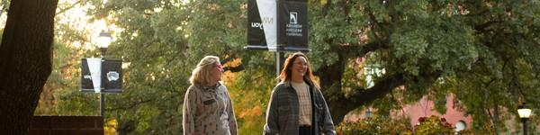 Two female students walk on campus with Old Main in the background.